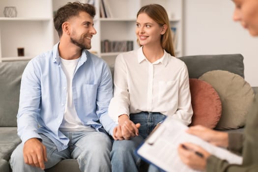 Cheerful millennial couple seated on therapist's couch sharing smiles