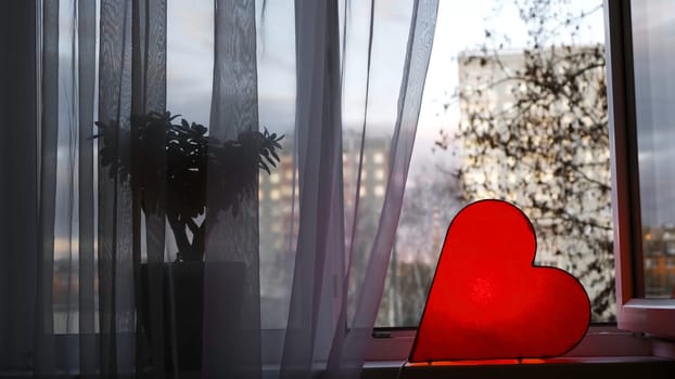 Bright lamp in shape of heart is on window. Concept. Red heart-shaped lamp stands at window and glows to bestow love on everyone. Bright romantic decor