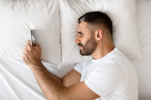 View from above of European guy using smartphone for internet communication, scrolling news and texting in social media applications while lying in bed indoors. Gadgets lifestyle