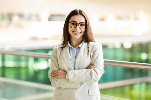 A professional young woman in smart attire and glasses stands with crossed arms