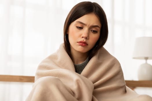 Upset young frozen woman wrapped with blanket sits on couch