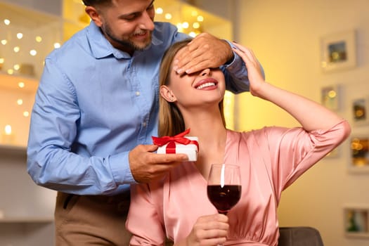 Man covering woman's eyes for a surprise with a gift, both with wine
