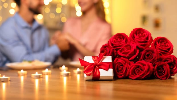 Gift box and roses on table with couple enjoying a romantic dinner