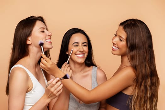 Smiling millennial international women in underwear apply nude makeup with brushes to friend