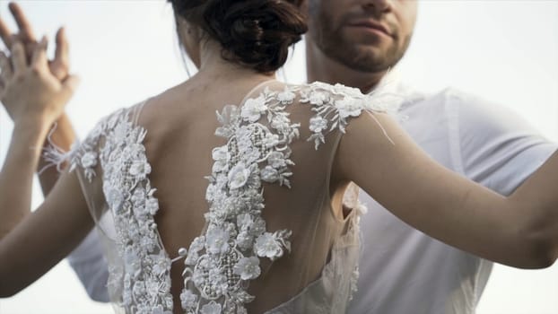 Rear view of a beautiful white wedding dress of a young slim bride with gentle flowers and cutout back. Action. Loving ang hugging bride and groom on bright sky background.