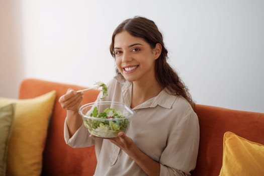 Young happy woman eating vegetable salad while sitting on couch at home