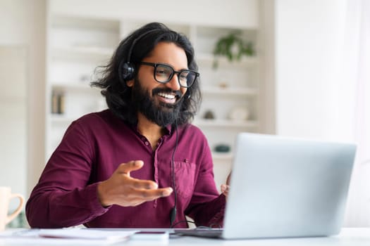 Cheerful indian freelancer wearing glasses and headset making video conference on laptop