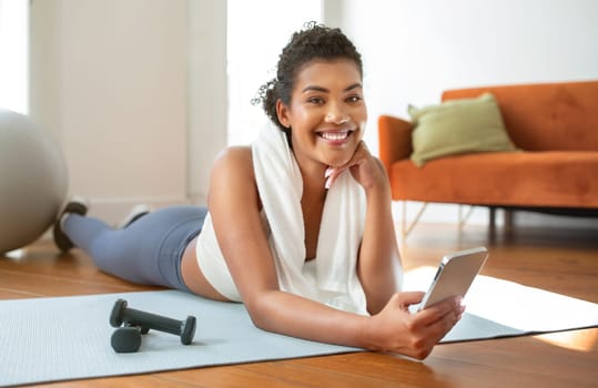 Sporty lady websurfing on mobile phone resting during domestic fitness workout, lying on gym mat in living room interior, smiling to camera posing with smartphone. Training and sport apps