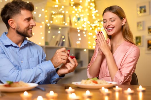 Man proposing to delighted woman with ring, festive lights in background