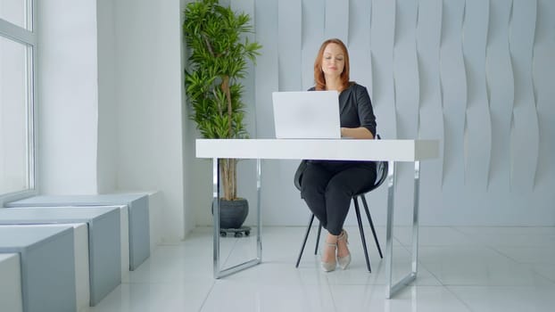 Woman in suit working in a modern office at the table. Action. White walls, green plant and a table with a laptop, minimalistic style.