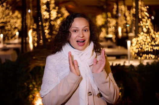 Beautiful woman expressing amazement standing against a city street illuminated with Christmas garlands at night time