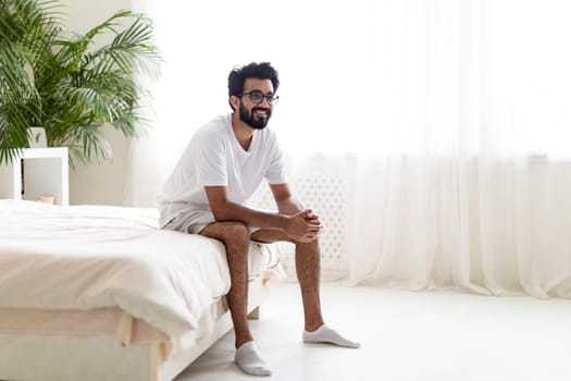 Happy Young Indian Man Sitting On Bed In Bedroom After Waking Up