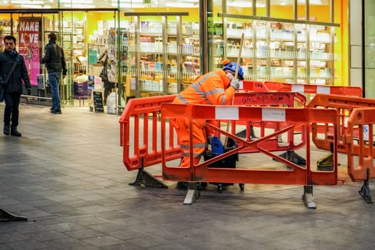 London, United Kingdom - February 01, 2019: Anonymous man wearing orange high visibility jacket cleaning floor at shopping centre, red barriers around him, light up stationery shop in background