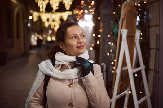 Authentic charming young woman smiles looking aside, admiring the Christmas decorations on the street in the night time