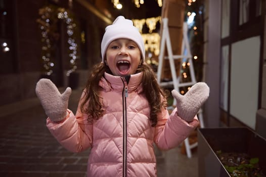 Emotional little girl smiles looking at camera, enjoying the Christmas atmosphere outdoors in the fairground at night