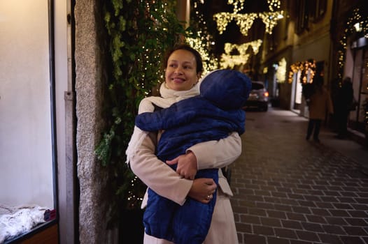 Happy loving mother carrying her baby, enjoying walk on the street in the night time, decorated with Christmas lights