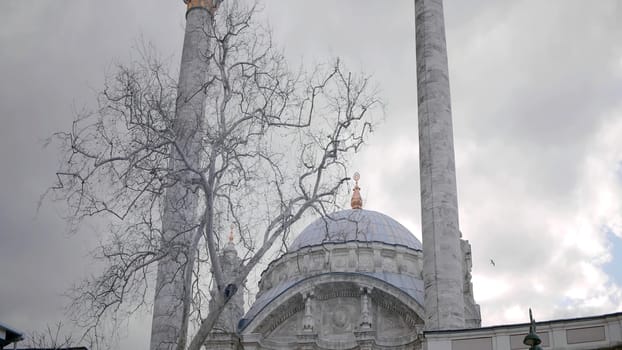 Mosque with minarets on background of cloudy sky. Action. Bare tree on background of mosque in cloudy weather. Beautiful white mosque with minarets in gray cloudy weather in autumn
