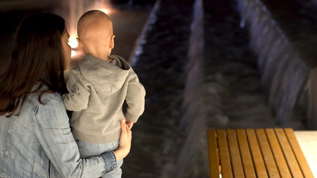 Rear view of young mother with her little son standing near the fountain in the late evening. Stock footage. Mom and her boy looking at the fountain.