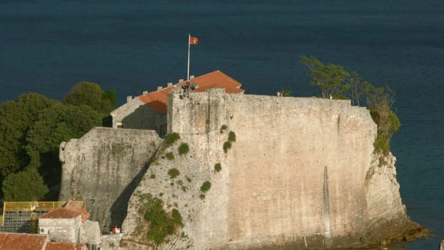 Montenegro, Budva - July 29, 2021: Ancient citadel fortress with red roofs. Creative. Stone fortress with rocky cliff by sea. Medieval citadel with stone walls and tourists near coast