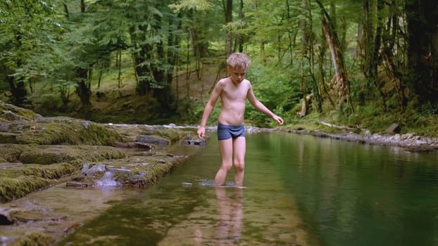 Boy walks on rocks in water in wild forest. Creative. Boy in swimming trunks walks on water in forest. Naked boy walks by water in green forest in summer