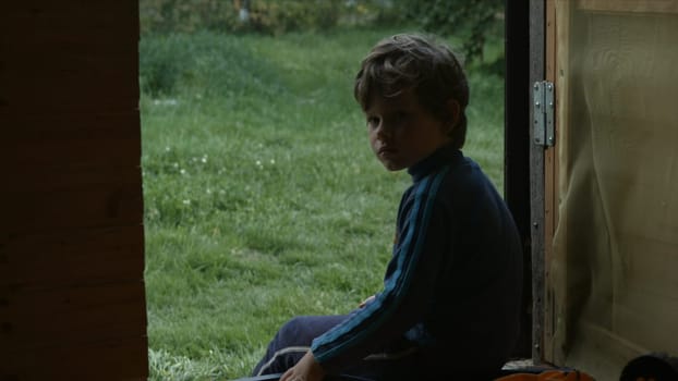 Beautiful boy is sitting on porch of house . Creative. Little boy is sitting in door on background of green grass. Beautiful Russian boy is resting in house in village.