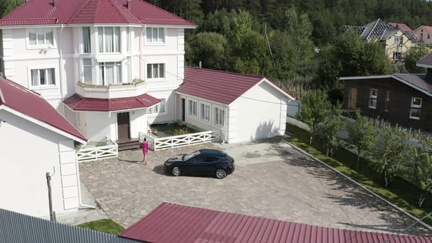 Aerial view of a woman in pink elegant dress coming home to her countryside cottage. Video. Business woman locking her expensive car and going towards beautiful three storey white house.