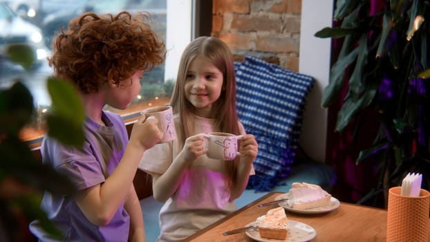 Two cute little kids having sweet meal in a cafe. Stock footage. Boy and girl holding cup of milk cocoa and looking happy.