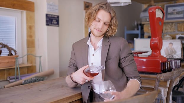 A handsome man with curly blond hair in beige suit pouring rose wine into a transparent glass. Action. Male sommelier pouring fruit wine at a bar.