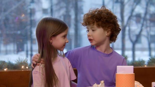 Children sitting together at a table in a restaurant. Stock footage. Brother with red curly hair putting ihs arm around the shoulders of her cute sister.