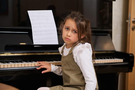 Little child girl pianist sitting at grand piano, feeling sadness and difficulties while learning playing grand piano