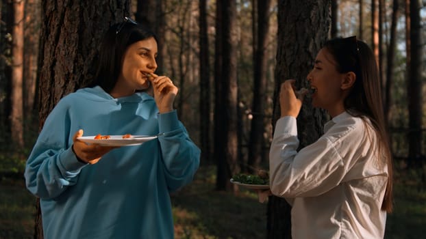 Female friends standing by the tree with plates and eating food while talking and smiling. Stock footage. Camping with friends in forest