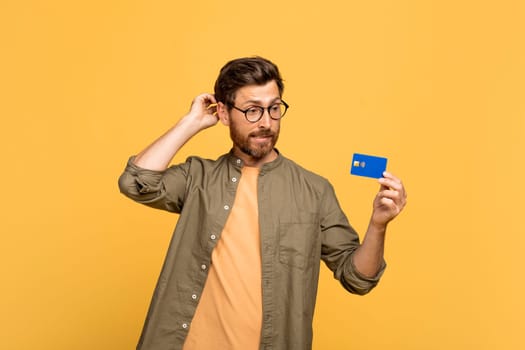Pensive middle aged man scratching head, look at credit card and think over purchases, yellow studio background