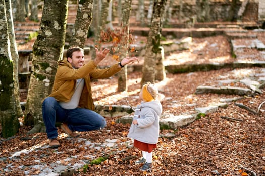 Smiling dad tosses fallen leaves over little girl standing in autumn park