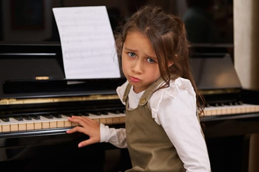 Emotional portrait of a cute little child girl, sitting at grand piano, expressing tiredness of learning a music lesson