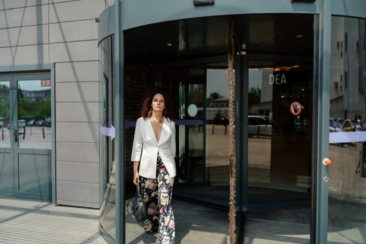Woman leaves a supermarket. Caucasian model with long dark hair, wearing a white jacket and colored trousers.