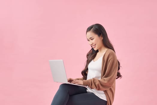 Beautiful young woman sitting on chair, working on laptop against pink wall