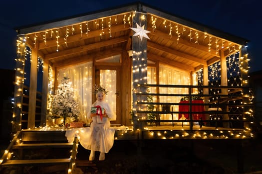 Happy blonde as Snow Maiden in white costume. Wooden house exterior Festive setting. Character with crown creating festive scene