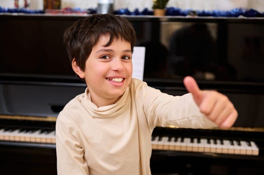 Cheerful elementary age schoolboy thumbing up and smiling looking at camera, sitting at piano during music lesson