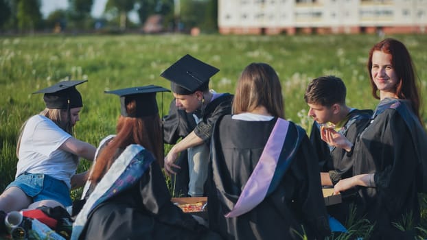 Graduates in black suits eating pizza in a city meadow.
