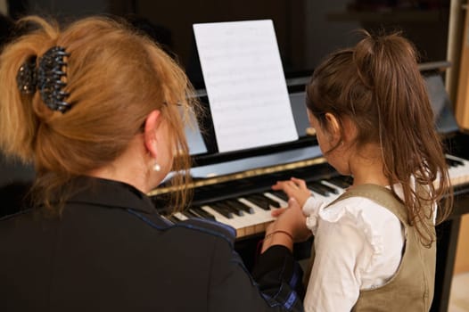 Rear view mature woman teacher musician and little child girl student playing together classic melody on old grand piano