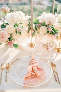 Knotted pink napkin lies on a plate on a set table near golden cutlery
