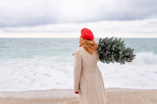 Redhead woman Christmas tree sea. Christmas portrait of a happy redhead woman walking along the beach and holding a Christmas tree on her shoulder. She is dressed in a light coat and a red beret.