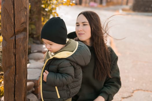 Cute little asian boy stands with millennial mother outdoors. Happy child walking in autumn park. Toddler baby boy wears trendy jacket and hat. Autumn fashion. Stylish child outside.