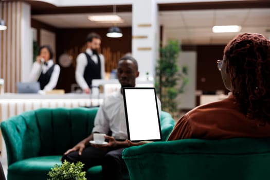 African american people at hotel using tablet with white screen, looking at blank template display and sitting in lounge area. Female guest holding device with isolated copyspace.