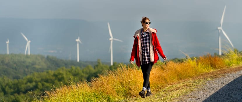 Caucasian woman with red coad and sunglasses walk along the roadside near the meadow and wind turbines or windmill with warm morning light.