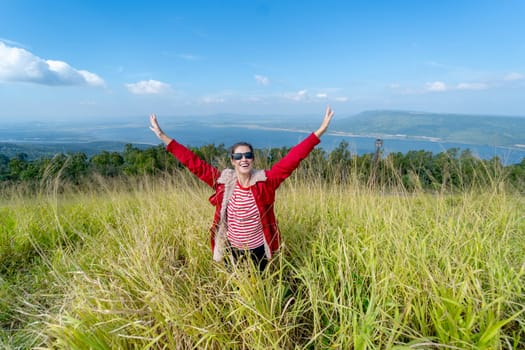 Caucasia woman with red coat and sunglasses spread her arms and stand in the meadow with background of water reservior and mountain.