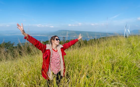 Wide shot of Caucasia woman with red coat and sunglasses spread her arms and stand in the meadow with background of water reservior, wind turbines and mountain.