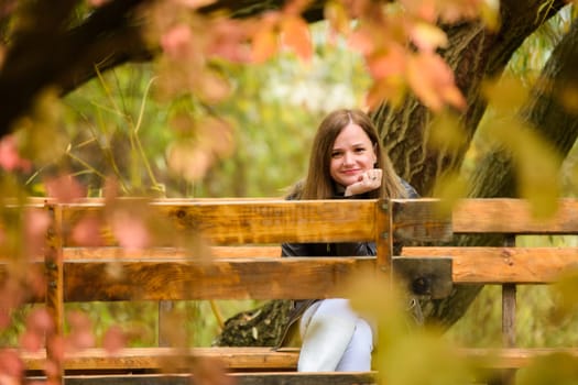 A young beautiful girl sits on a bench in an autumn park and looks into the frame, smiling
