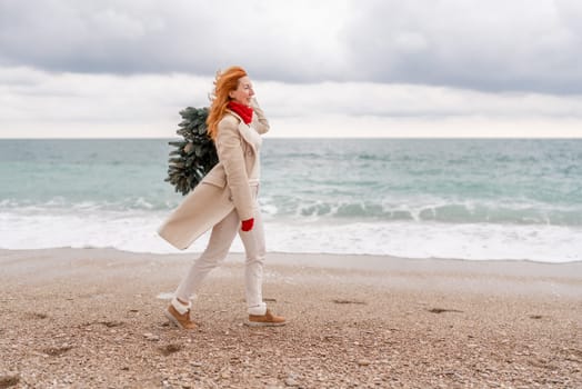 Redhead woman Christmas tree sea. Christmas portrait of a happy redhead woman walking along the beach and holding a Christmas tree in her hands. She is dressed in a light coat and a red beret.