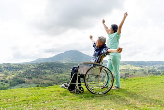 Wide shot of Asian nurse and senior man on wheelchair action of stretching together and both stay near the cliff with mountain and village as background.